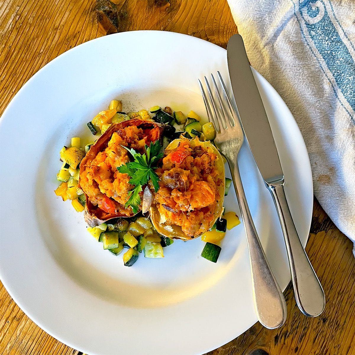 A white plate with stuffed peppers filled with a vegetable and potato mixture, garnished with parsley, and surrounded by diced sautéed courgette. A fork and knife rest at the side of the plate, on a wooden table.