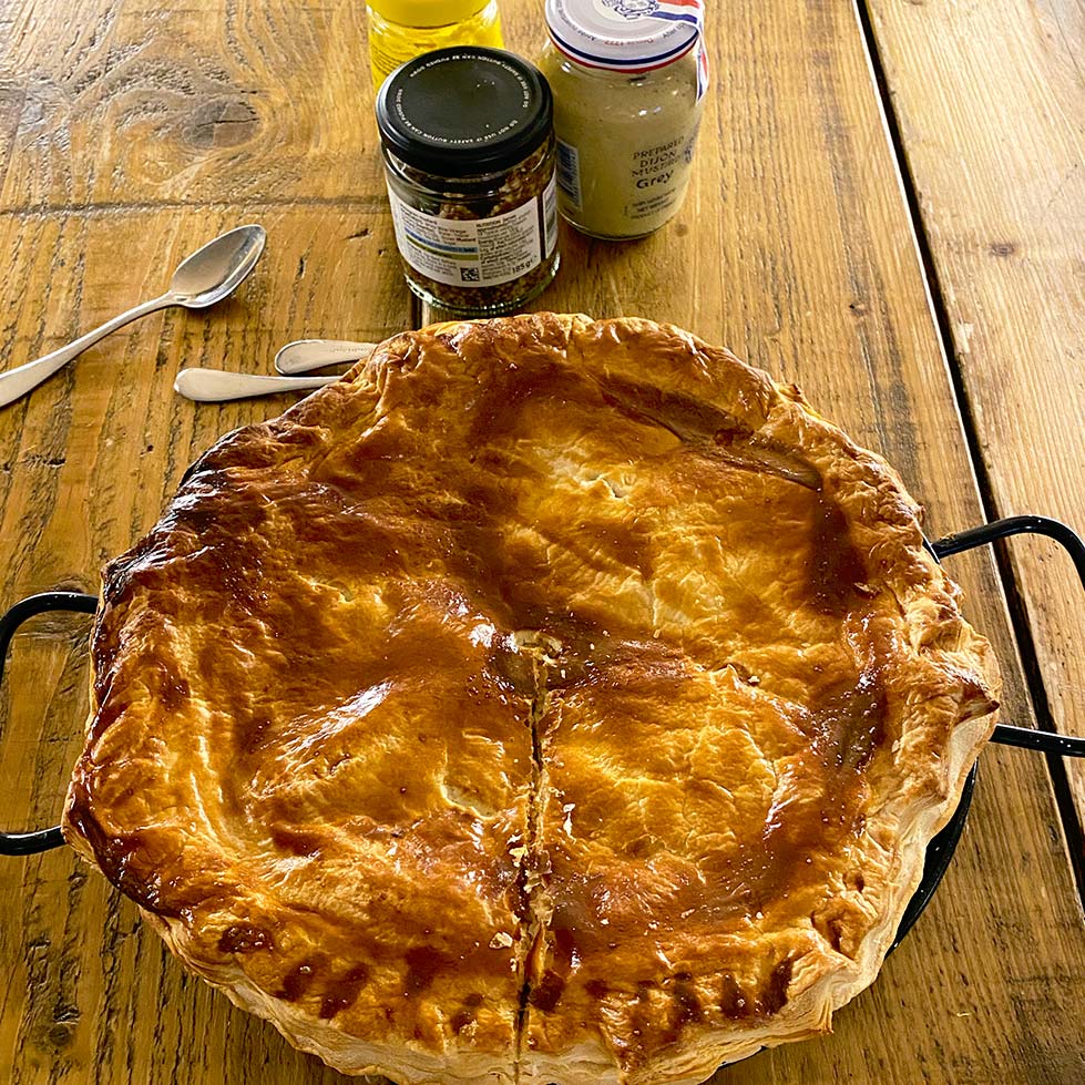 A golden-brown, freshly baked pie in a tin sits on a rustic wooden table, with two spoons and jars of condiments in the background. The pie has a flaky, glossy pastry lid and a single cut starting from the centre.