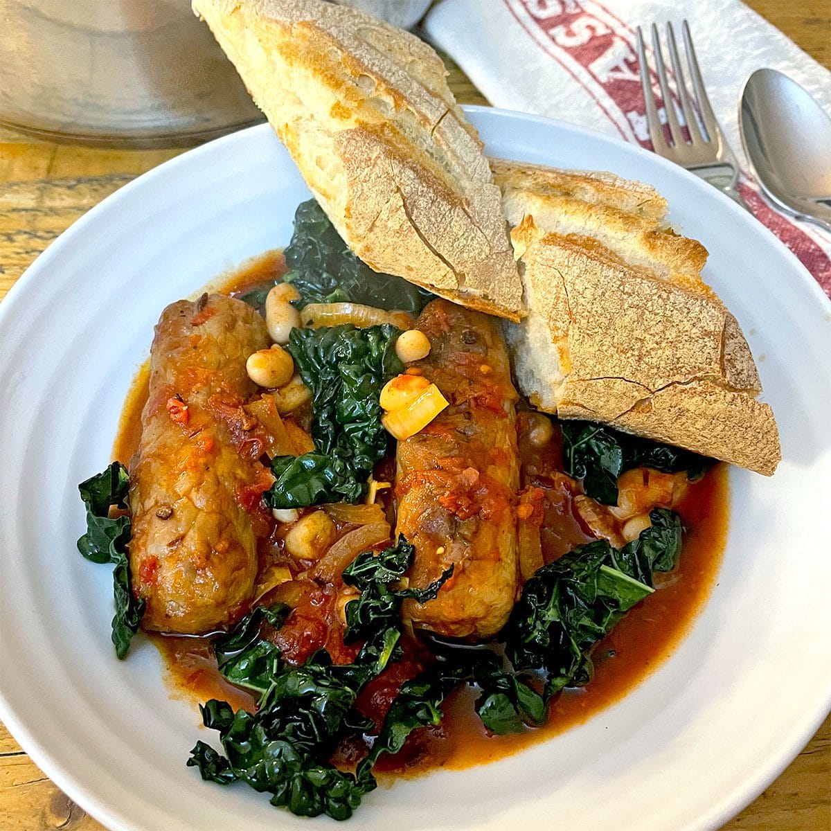 A white plate with sausage stew, featuring sausages, leafy greens, beans, and tomato sauce, garnished with two pieces of crusty bread on top. A fork and spoon rest on a serviette nearby.