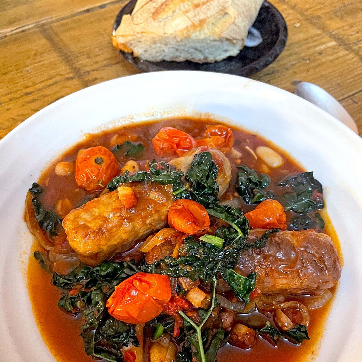A white bowl filled with sausage stew, featuring sausages, cherry tomatoes, greens, and beans in a tomato-based sauce. A crusty piece of bread sits on a small black plate in the background.