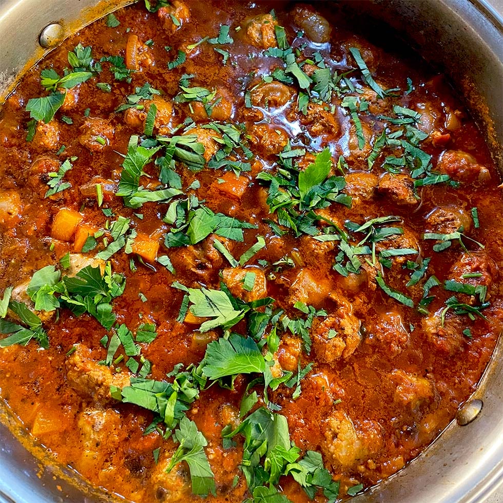A close-up of a pot filled with a chunky tomato-based stew, featuring pieces of meat, vegetables, and topped with fresh chopped herbs.