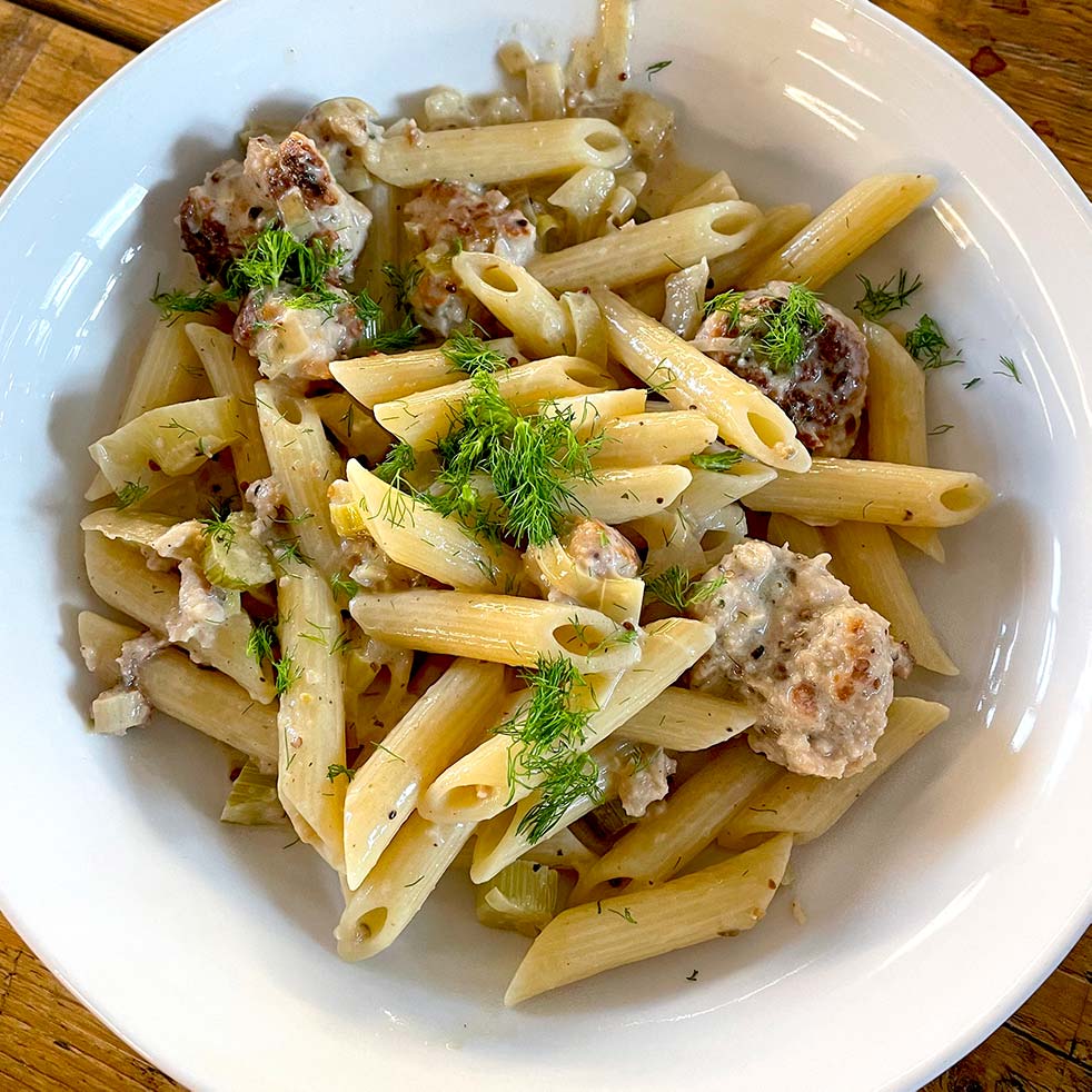 A white bowl filled with penne pasta, meatballs, creamy sauce, and garnished with fresh herbs, sitting on a wooden table.