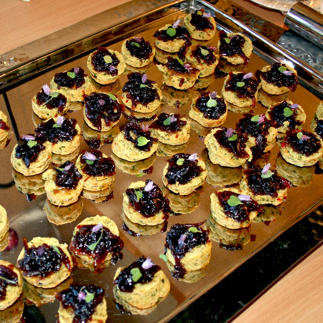 A tray filled with small, round appetiser bites topped with a dark jam or chutney and garnished with tiny herbs and edible flowers, arranged neatly for serving.