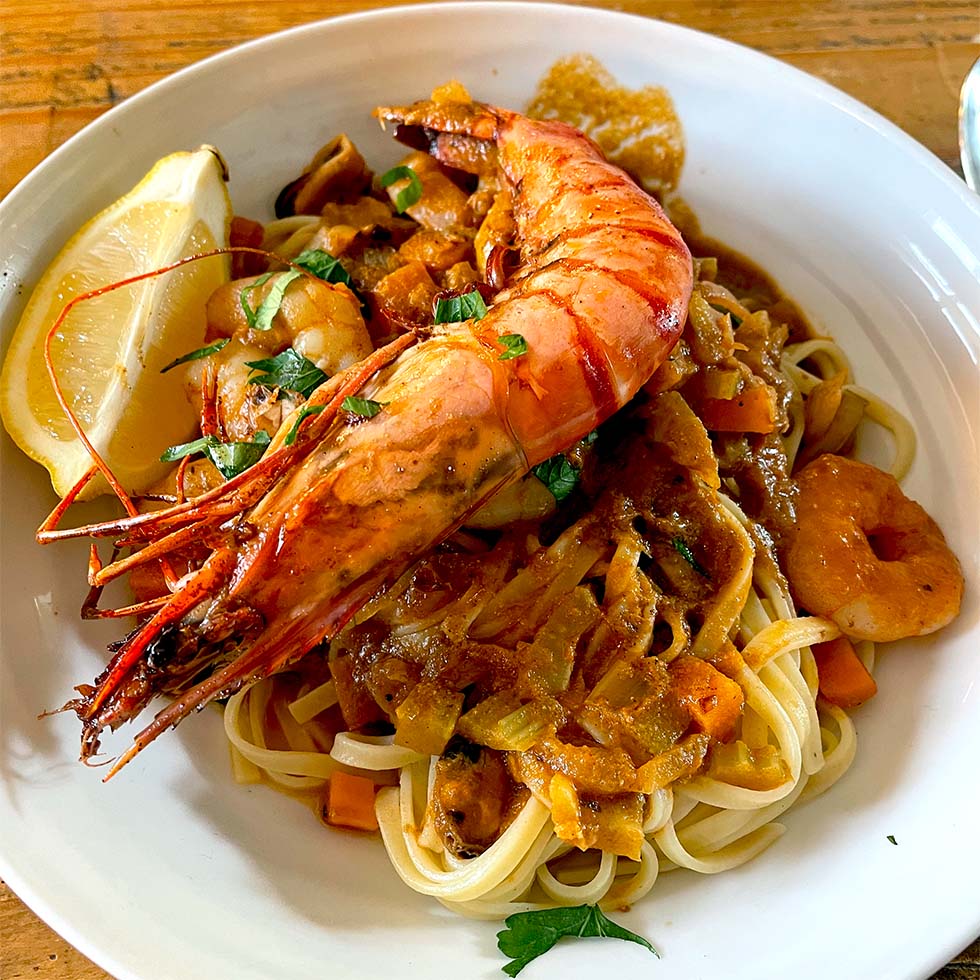 A bowl of pasta topped with a large grilled king prawn, creamy tomato sauce, chopped herbs, and a lemon wedge on the side. Prawn pieces and diced vegetables are mixed in with the pasta.