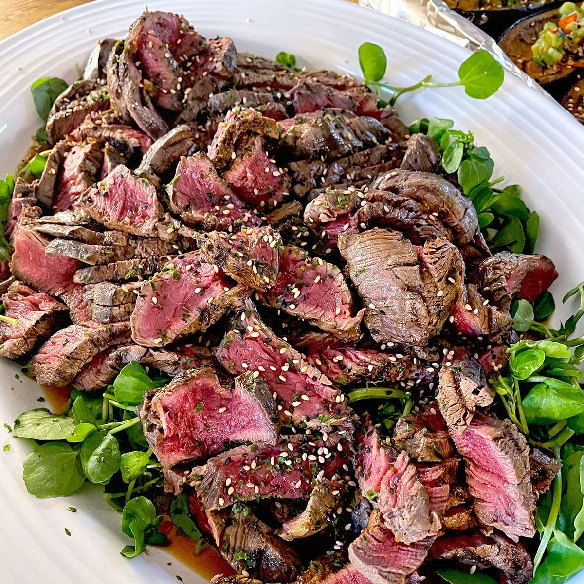 Slices of medium-rare steak topped with sesame seeds, served on a bed of fresh salad leaves on a white platter.