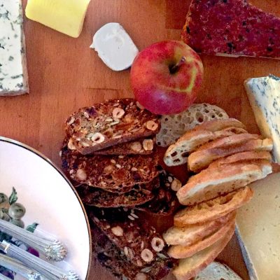 A wooden table with slices of baguette, nutty crispbread, an apple, various cheeses, and a plate with cutlery in the corner.