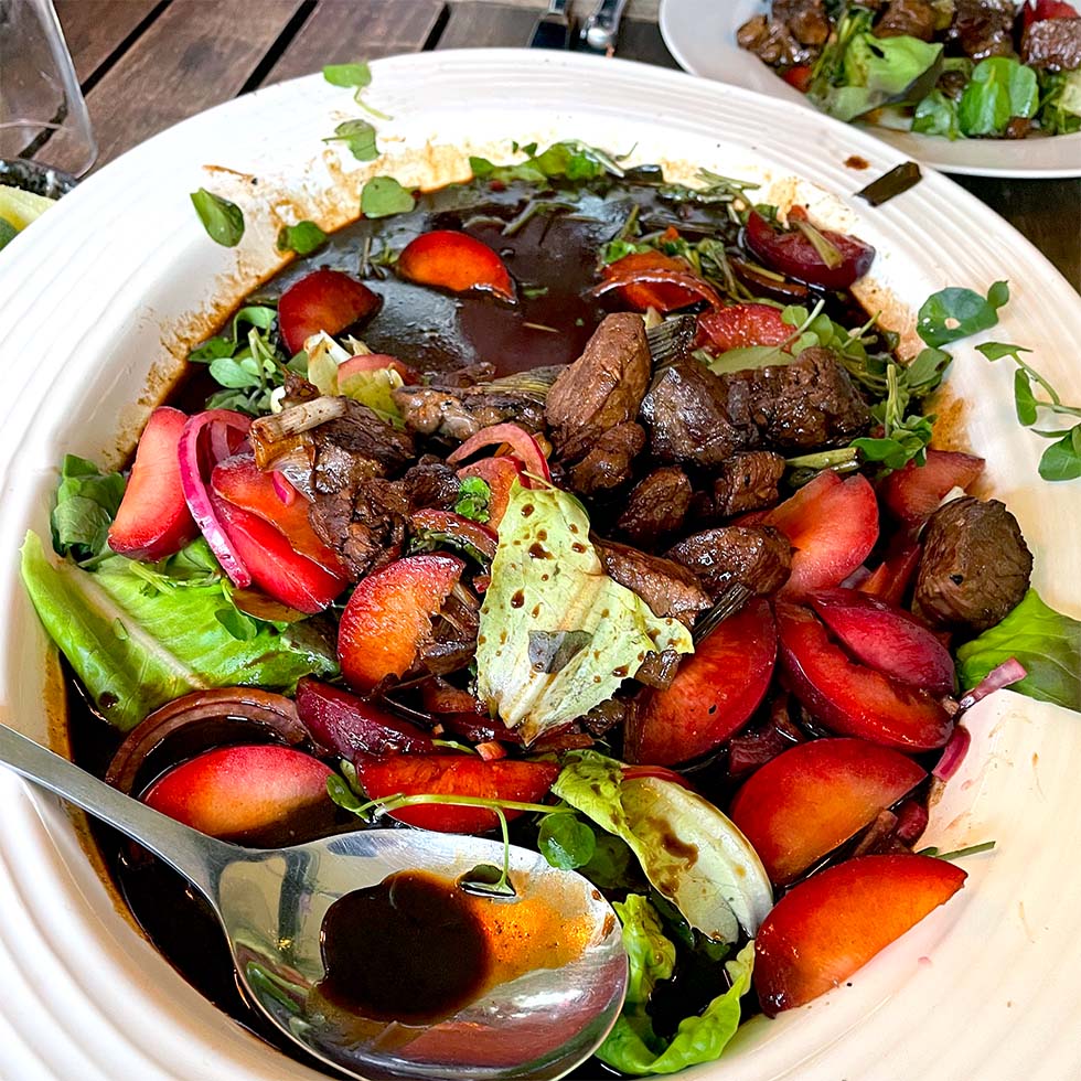 A white bowl filled with a colourful salad of leafy greens, sliced plums, seared beef pieces, and a dark balsamic dressing, with a large serving spoon resting inside the bowl.