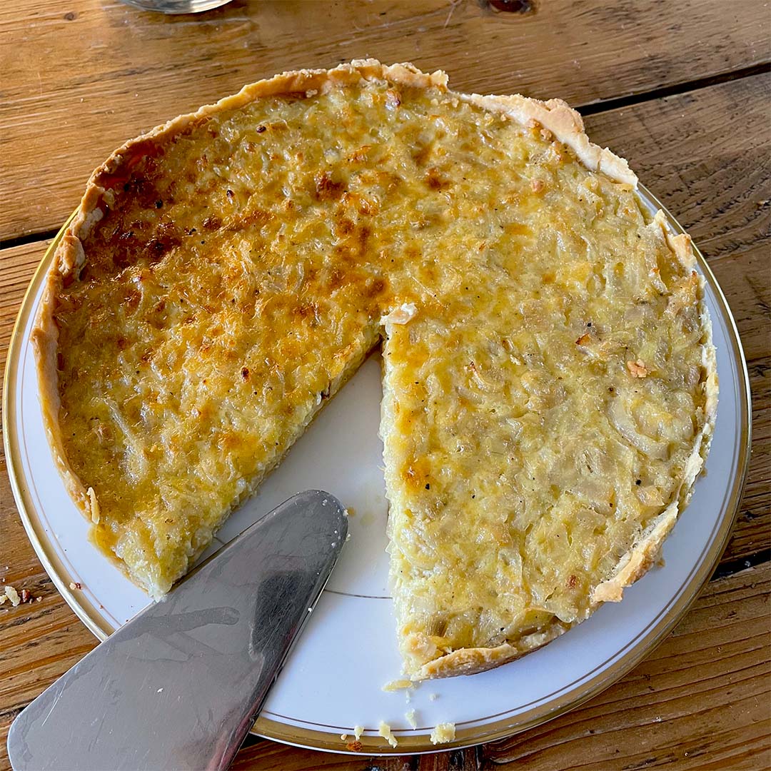 A partially sliced onion tart sits on a white plate atop a wooden table, with a metal serving spatula resting nearby. The tart has a golden-brown, slightly uneven surface and a flaky pastry.
