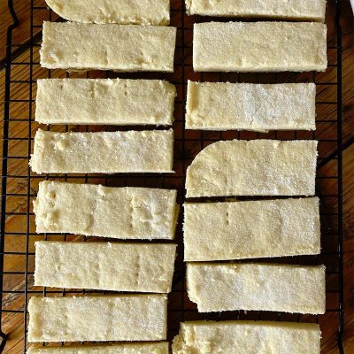 Rectangular pieces of golden, sugar-dusted shortbread are cooling on a black wire rack set over a wooden surface. The shortbread bars are arranged neatly in rows.