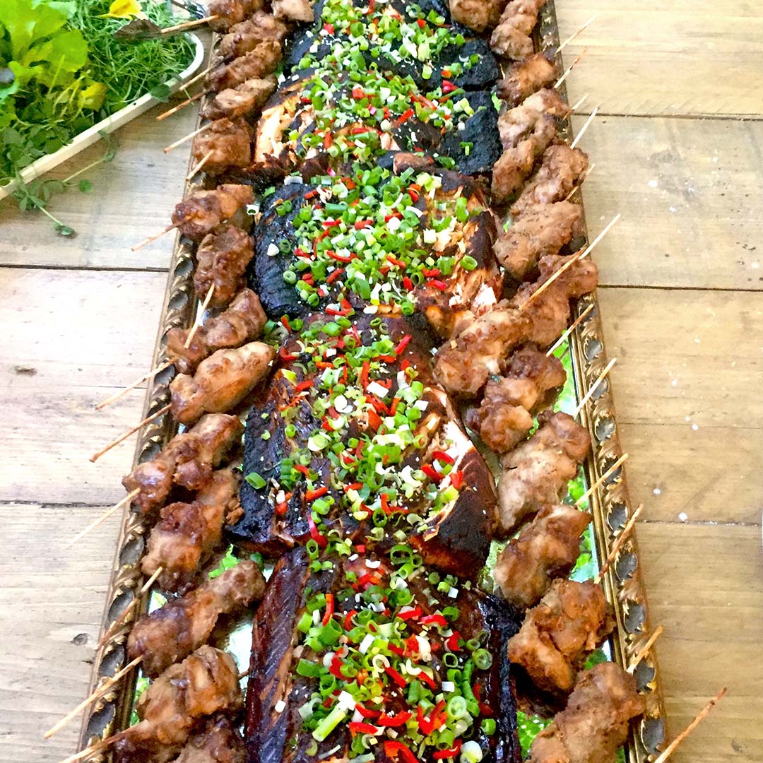 A large tray of grilled meat skewers arranged along the sides, with assorted grilled meats in the centre topped with chopped spring onions and red chillies, displayed on a wooden table.