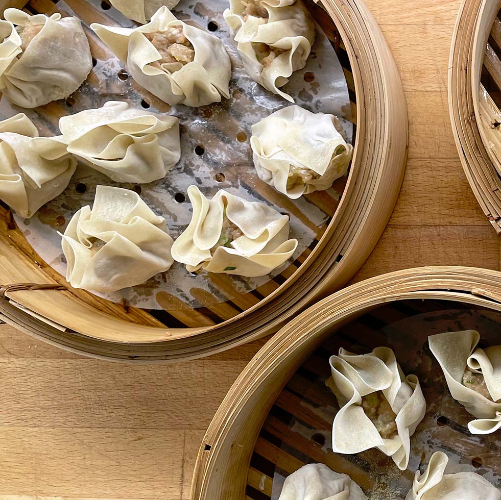 Three bamboo steamers filled with uncooked dumplings, arranged on a wooden surface. The dumplings are wrapped in thin pastry and placed on greaseproof paper inside the steamers.