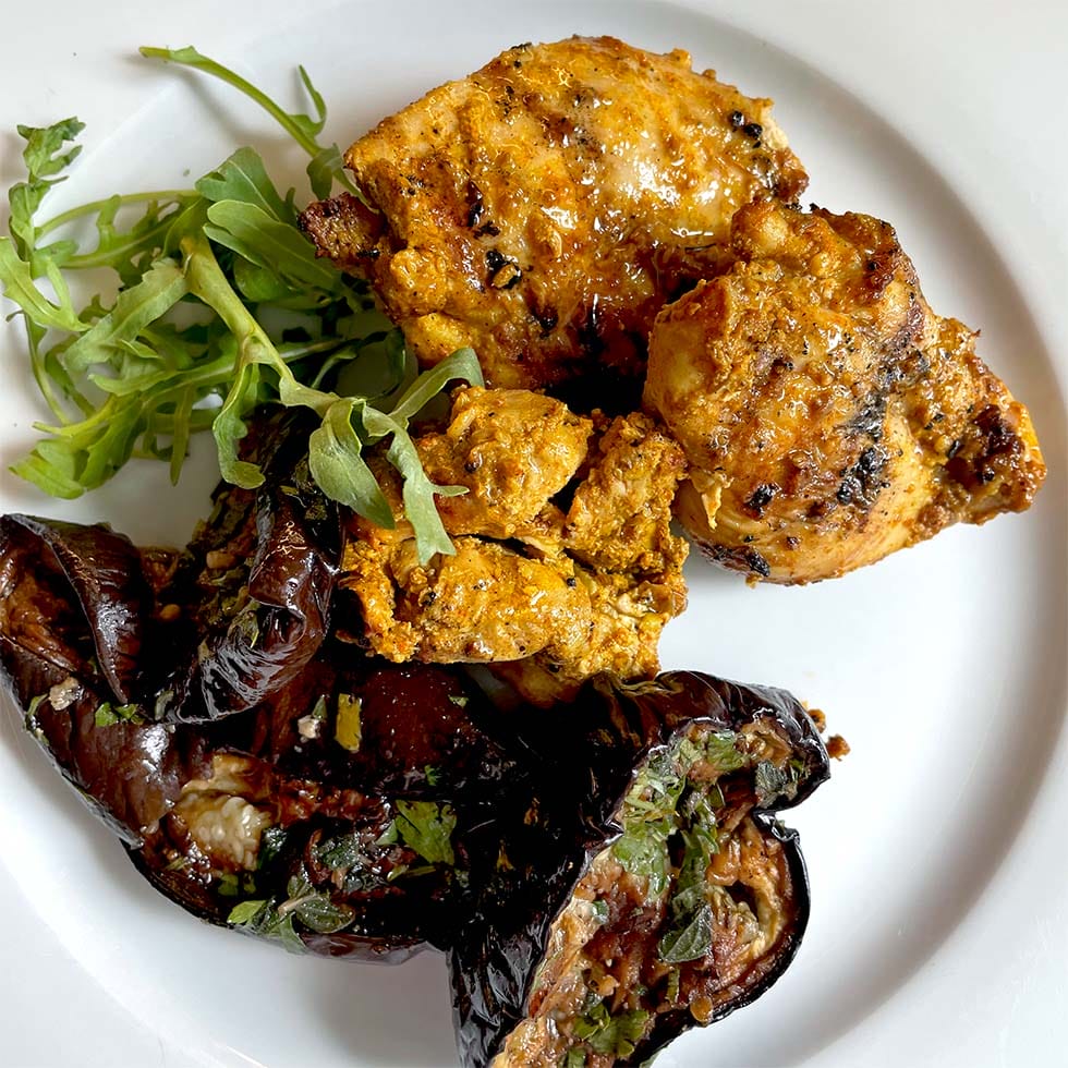 Grilled pieces of seasoned chicken served with roasted aubergine garnished with herbs and a small side of fresh rocket on a white plate.