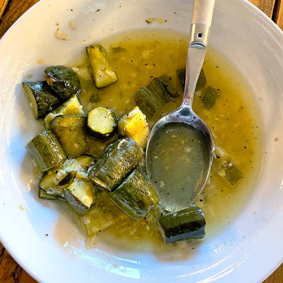 A white bowl containing chunks of cooked courgette in a yellow-green broth, with a metal spoon resting in the bowl. Some broth and vegetable pieces are scattered around.