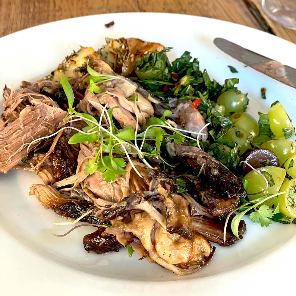A plate of shredded cooked meat with microgreens, roasted vegetables, sliced green grapes, and fresh herbs, served on a white dish on a wooden table. A knife rests on the plate’s edge.