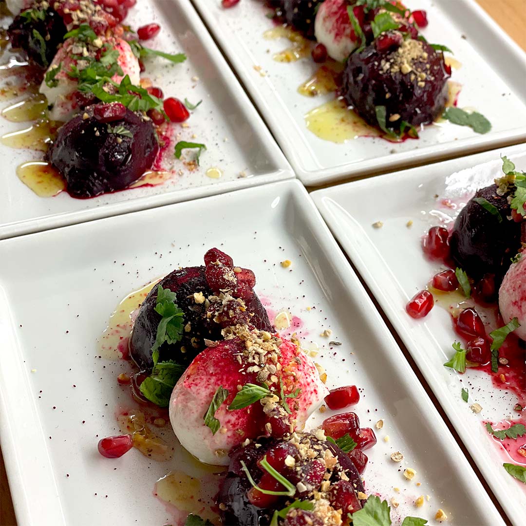 Four rectangular plates each with a vibrant salad of roasted beetroot, creamy cheese, pomegranate seeds, herbs, crushed nuts, and a drizzle of dressing, artfully arranged for serving.