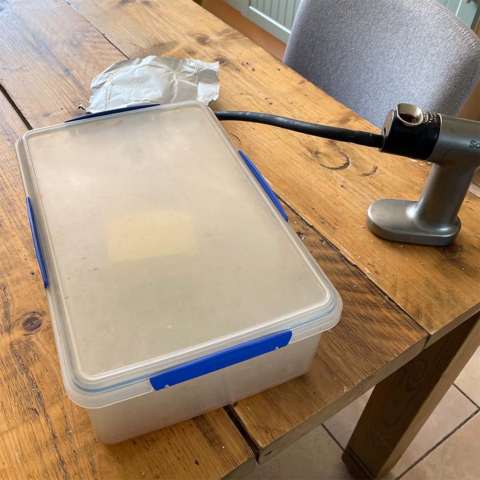 A plastic container with a blue catch sits on a wooden table beside a silver handheld kitchen tool with a black hose attached. A piece of crumpled paper is also on the table near the container.