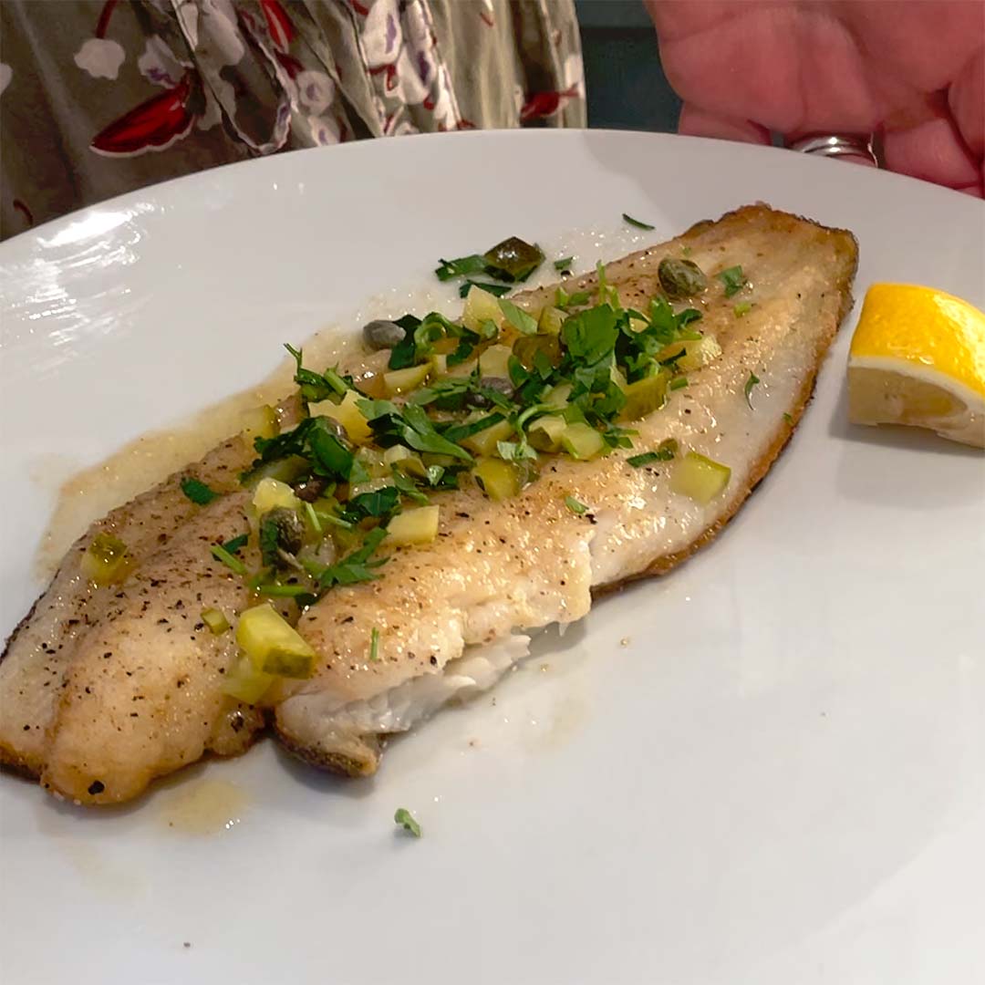 A cooked fish fillet garnished with chopped herbs and capers is served on a white plate with a lemon wedge on the side. A hand holds the plate, and part of a patterned garment is visible in the background.