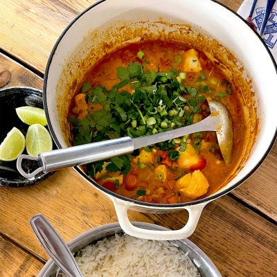 A pot of fish stew with chunks of fish, vegetables, and fresh herbs sits on a wooden table next to a bowl of white rice and a plate with lime wedges. A ladle rests inside the pot.
