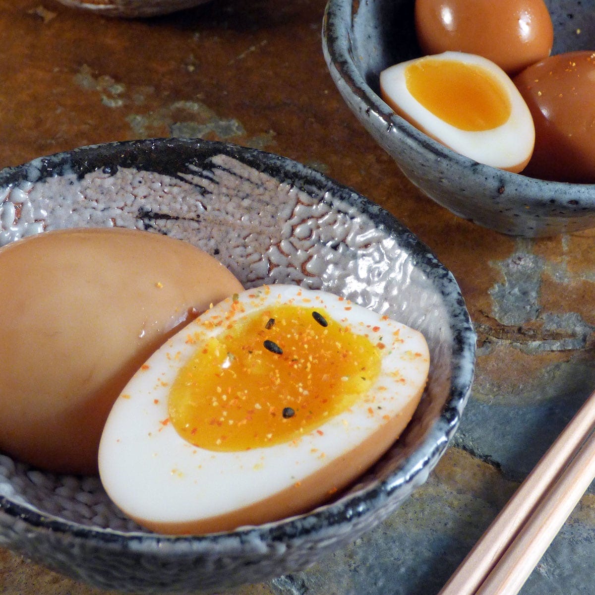 A ceramic bowl with a halved soft-boiled egg, seasoned with spices and black sesame seeds. Another bowl contains whole and halved eggs. Chopsticks are placed nearby on a stone surface.