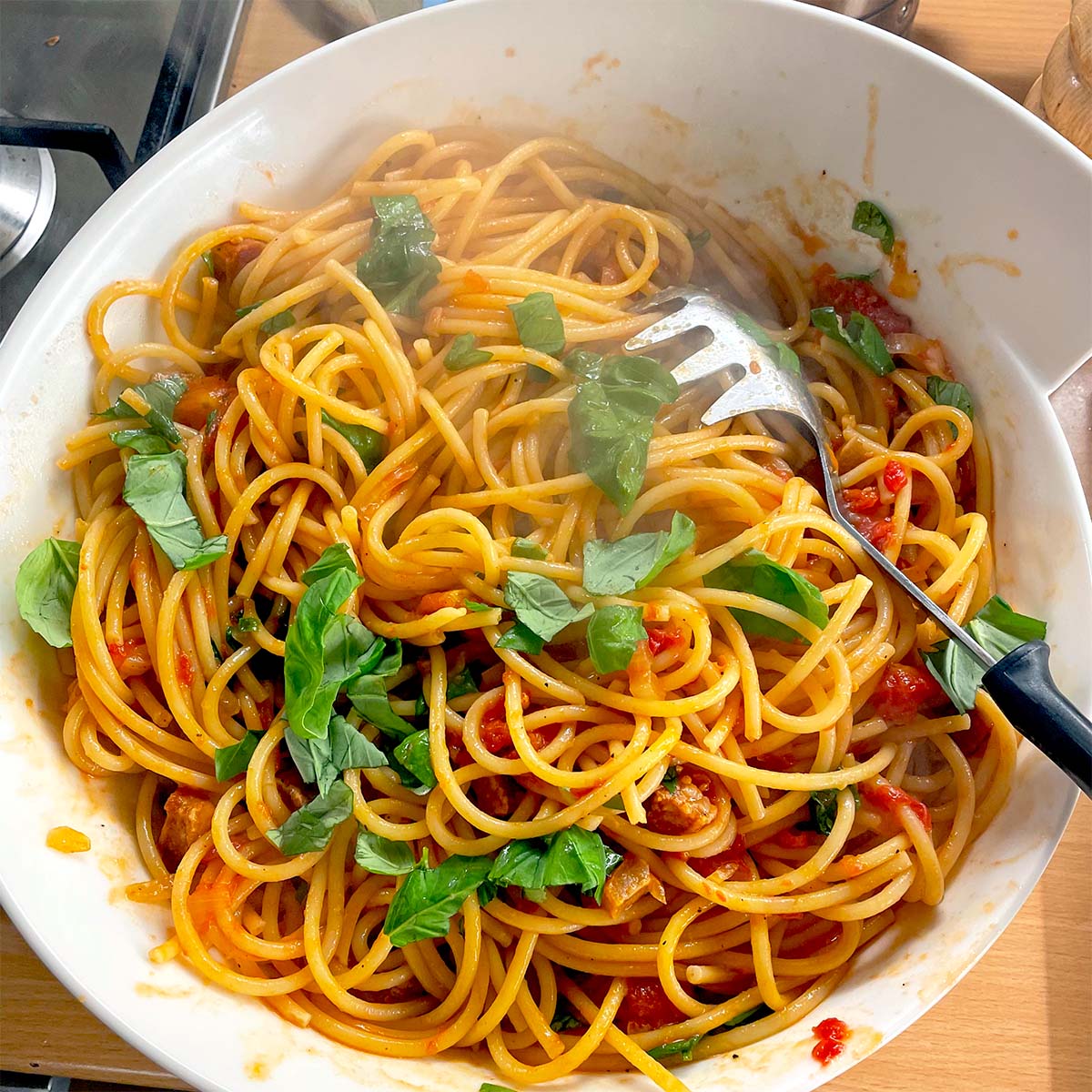 A bowl of spaghetti with tomato sauce, fresh basil leaves, and a pasta serving spoon, sitting on a wooden surface. Steam rises from the pasta, indicating it is freshly cooked.