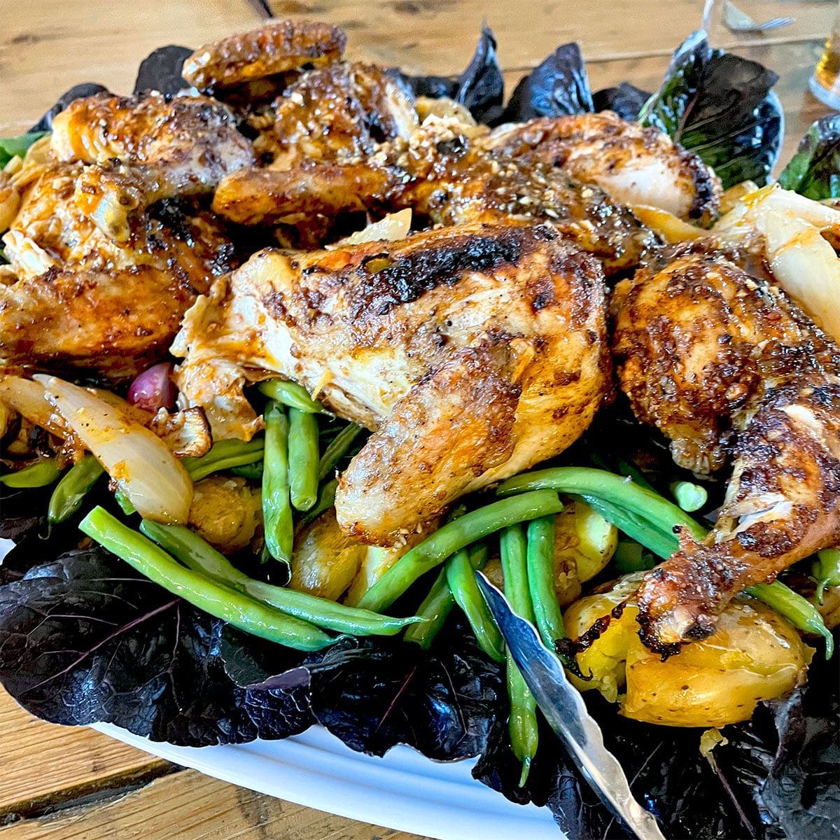 A plate of roast chicken pieces served on a bed of green beans, new potatoes, onions, and leafy greens, garnished with herbs.