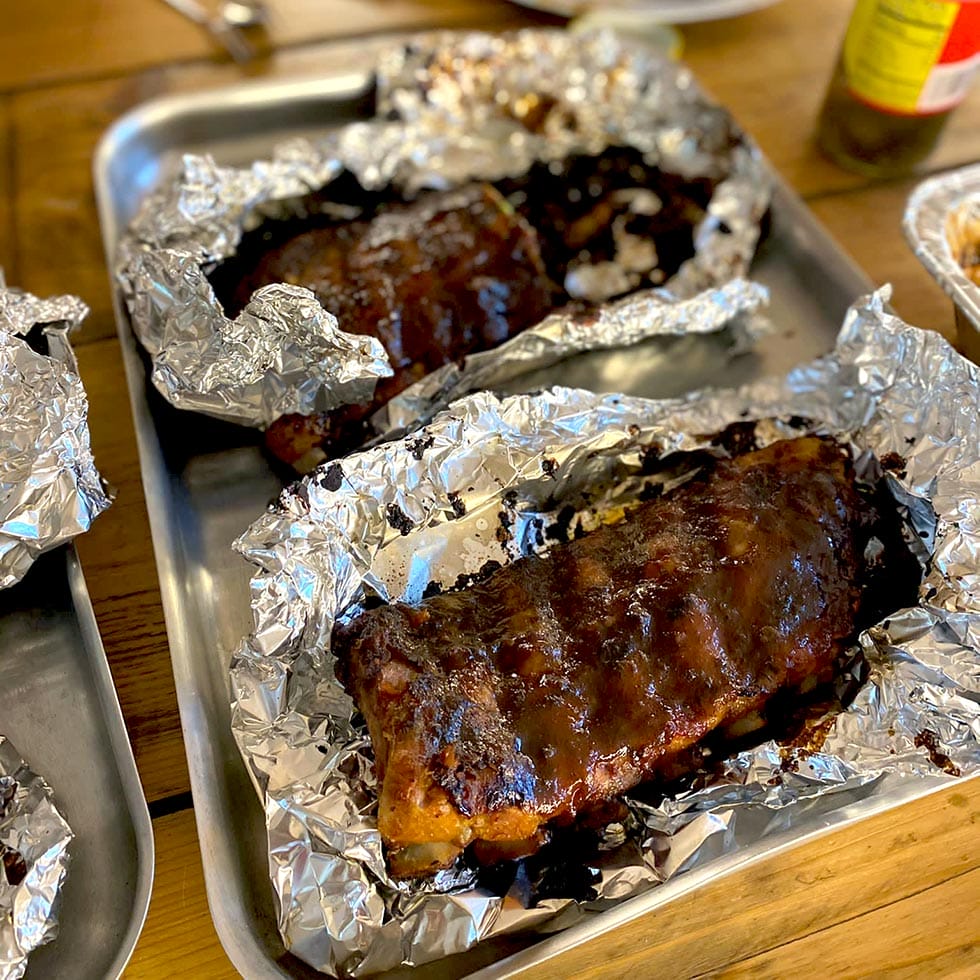 Barbecued ribs covered in sauce, partially wrapped in aluminium foil, served on metal trays on a wooden table. The ribs appear juicy and well-cooked, ready to be eaten.