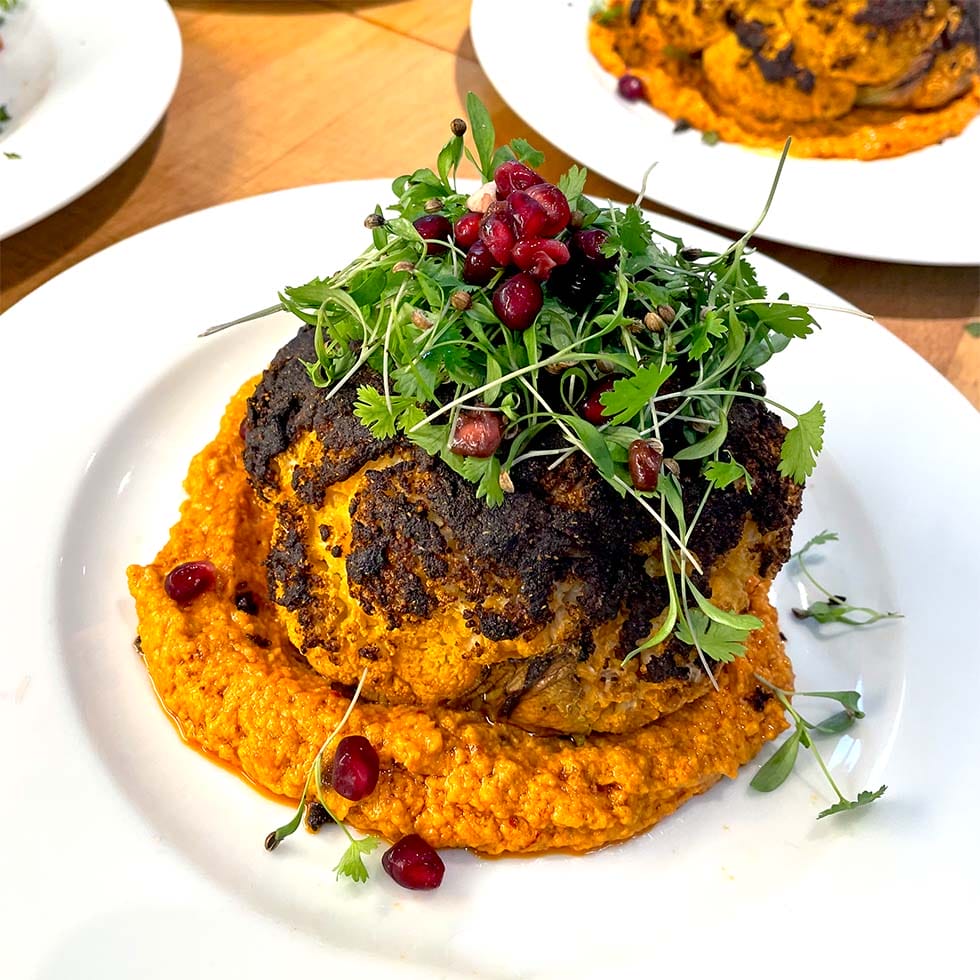 A whole roasted cauliflower sits on a bed of orange sauce, garnished with fresh microgreens and pomegranate seeds, served on a white plate.