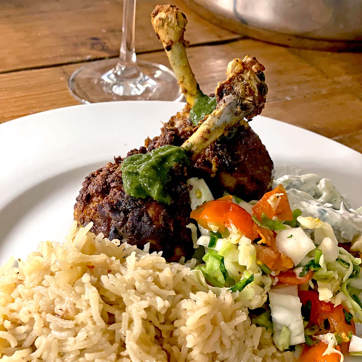 A plate of spiced chicken drumsticks topped with green sauce, served with white rice, chopped vegetable salad, and a side of creamy sauce, with a wine glass in the background.