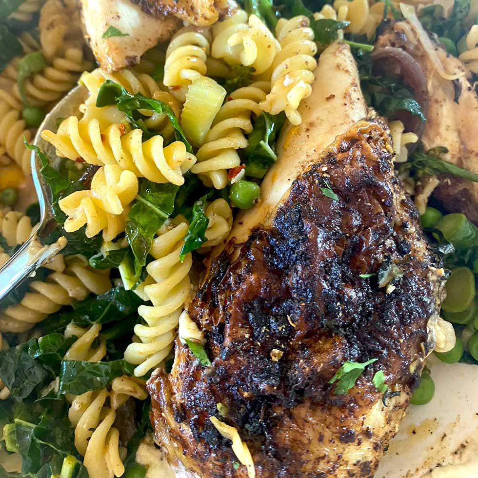 Close-up of a plate with seasoned grilled chicken breast, rotini pasta, leafy greens, peas, and vegetables, all mixed together. A fork is partially visible on the left side of the dish.