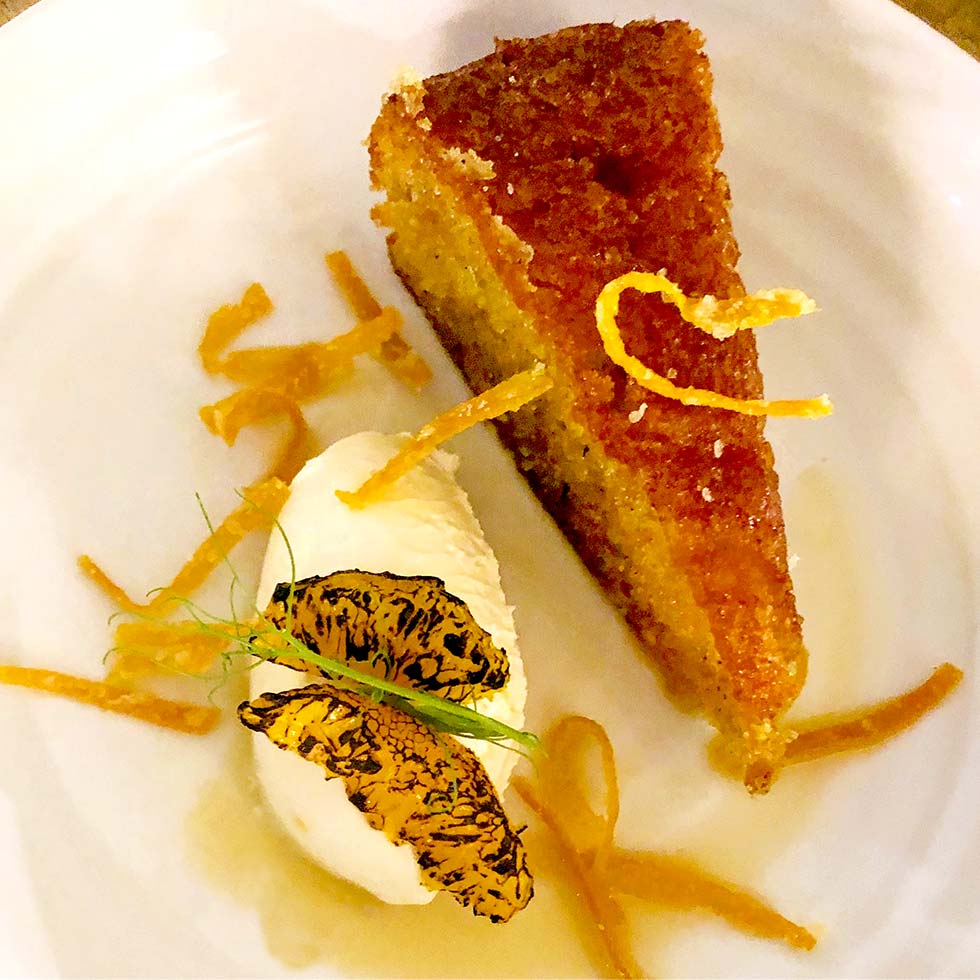 A slice of golden syrup cake garnished with thin orange zest strips, served with a scoop of vanilla ice cream and caramelised orange slices on a white plate.