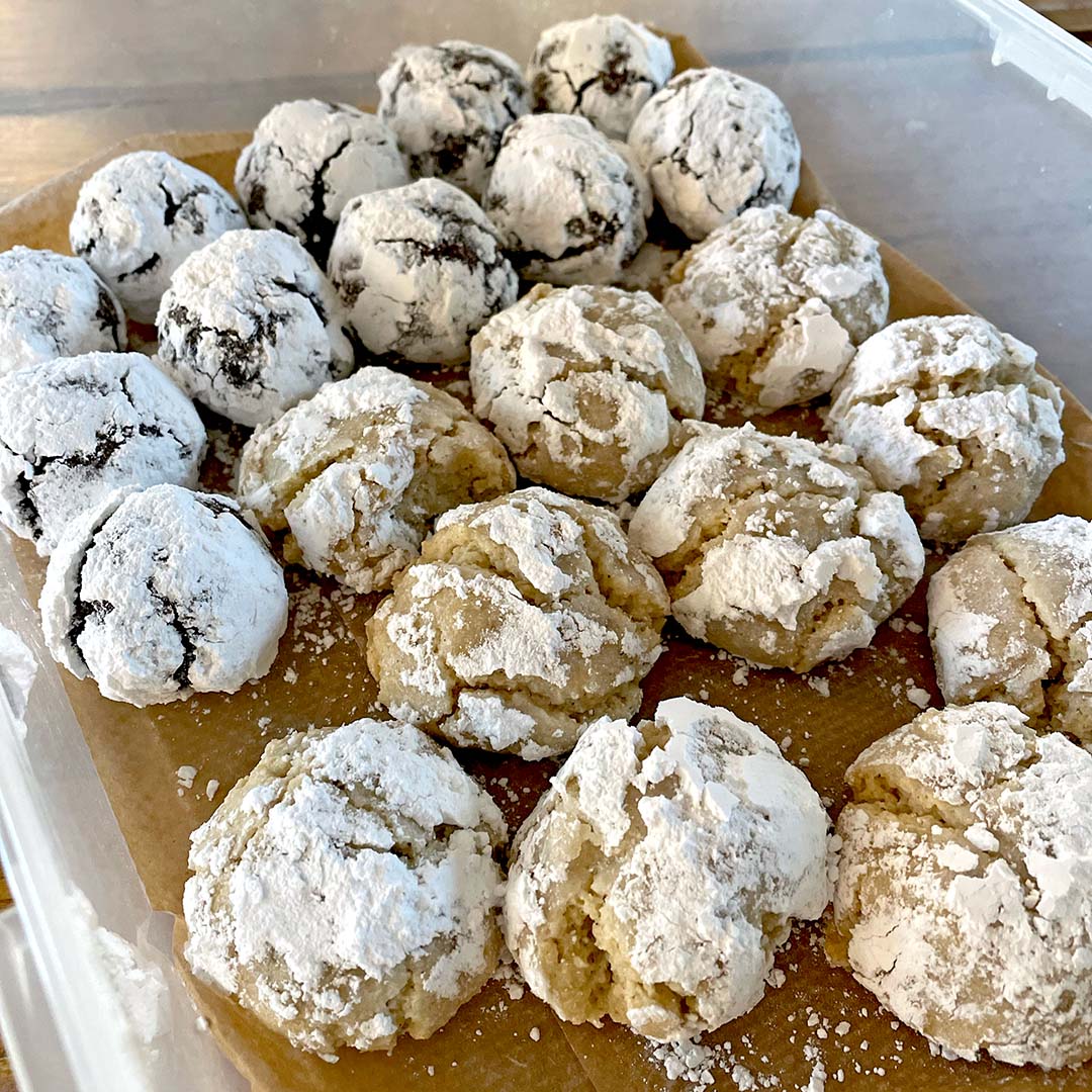 A container lined with brown baking parchment holds rows of crinkle biscuits. The biscuits are covered in icing sugar; some are chocolate and others are light-coloured, suggesting different flavours.