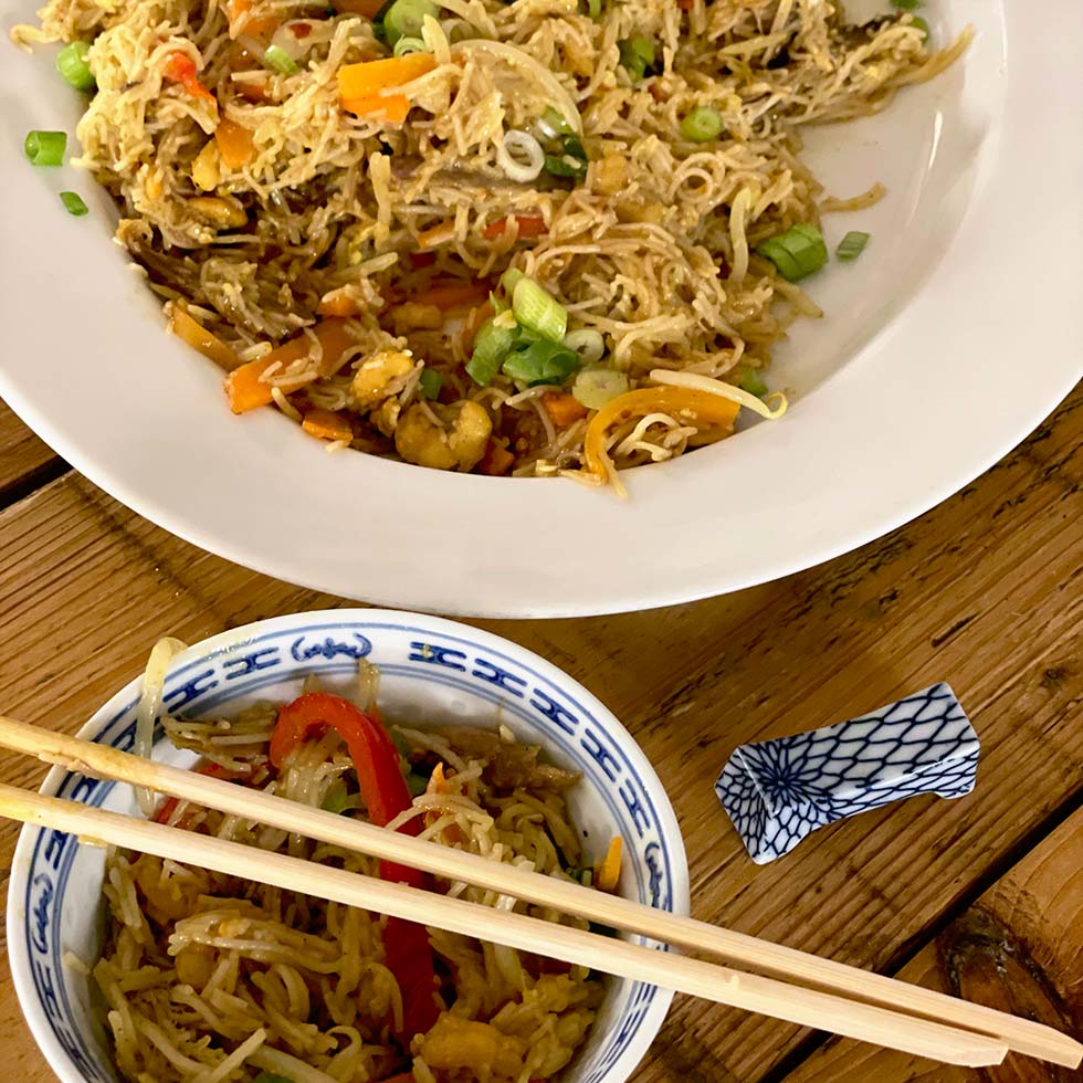 A large white plate and a small patterned bowl filled with stir-fried noodles, vegetables, and spring onions sit on a wooden table. Chopsticks rest on the bowl, with a chopstick holder nearby.