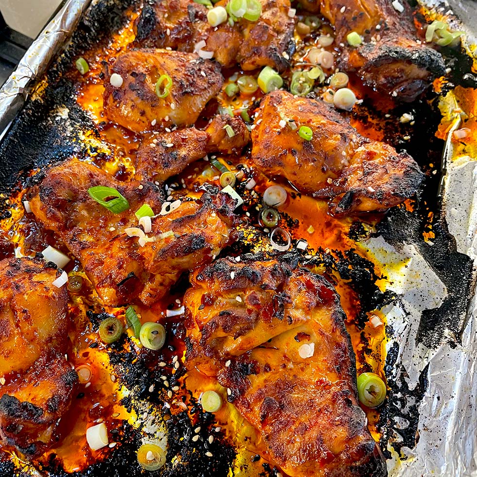 Charred spicy baked chicken wings on a foil-lined tray, garnished with sliced spring onions and sesame seeds. The wings are coated in a red sauce, with crispy edges and visible bits of seasoning.