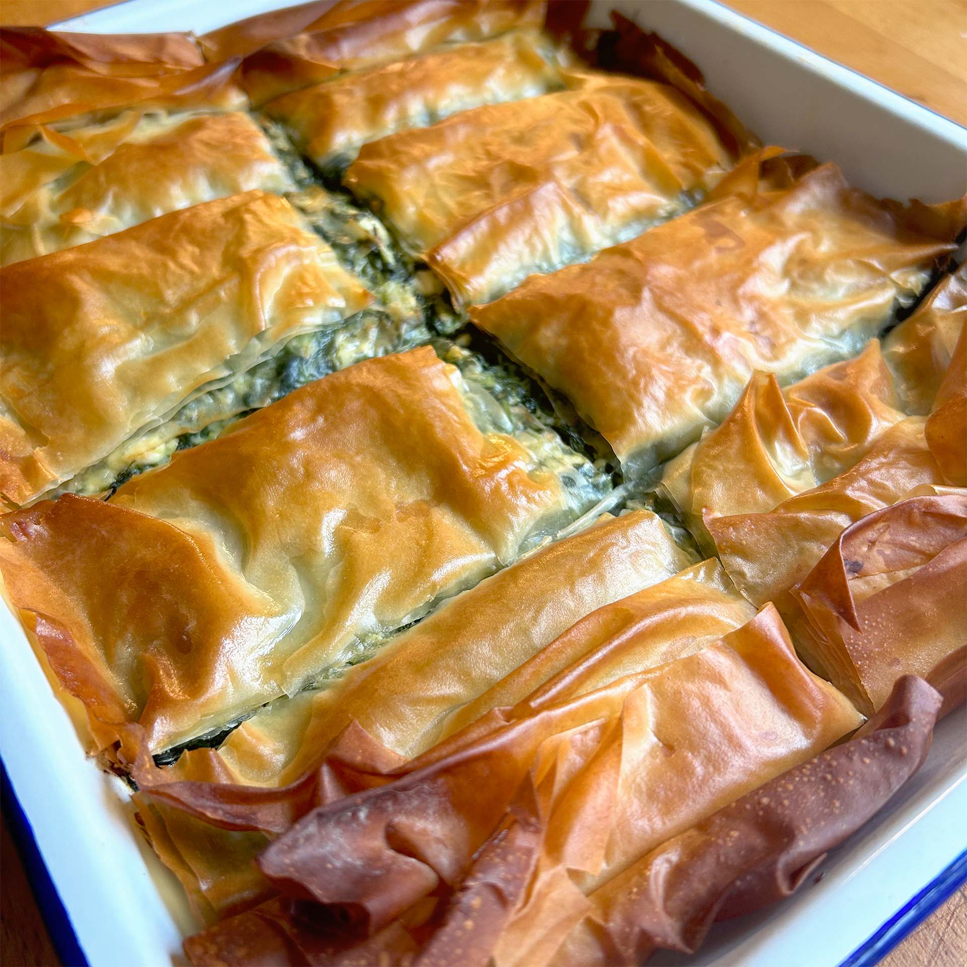A close-up of golden-brown, flaky spanakopita cut into rectangles in a white baking dish, with crispy filo pastry visible on top and edges.