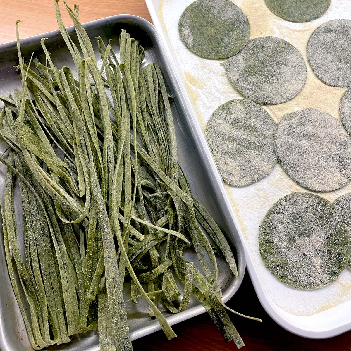 Fresh green pasta dough cut into fettuccine strips sits in one tray, while round green ravioli wrappers dusted with flour are arranged on another tray. Both trays are on a wooden surface.
