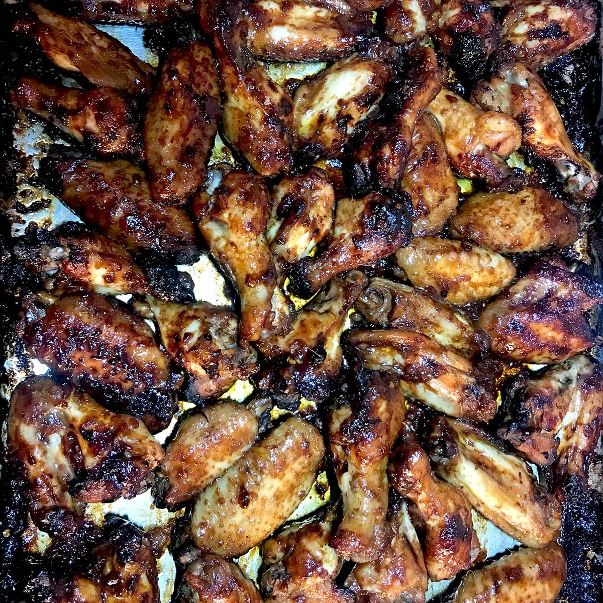 A close-up view of a baking tray filled with numerous baked chicken wings, golden-brown and caramelised, with crispy, charred edges.