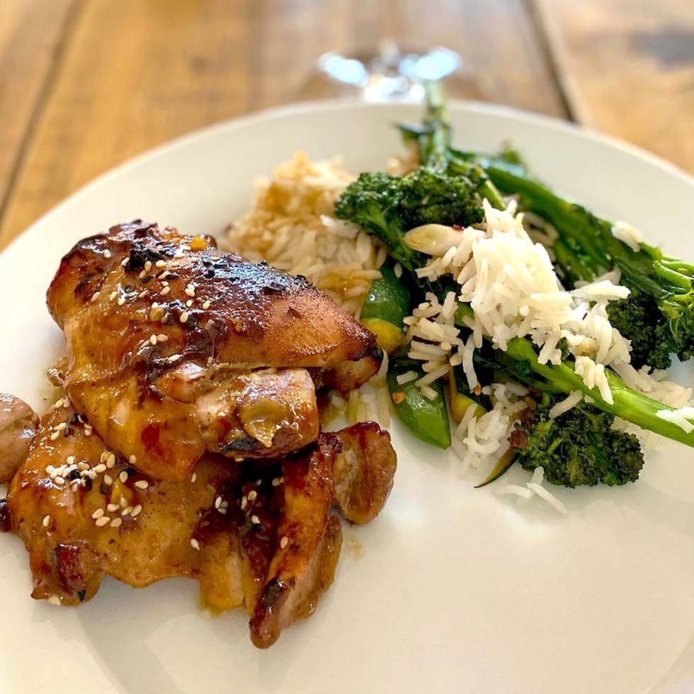 A plate of glazed chicken thighs sprinkled with sesame seeds, served with white rice and sautéed green vegetables, including tenderstem broccoli, on a white plate on a wooden table.