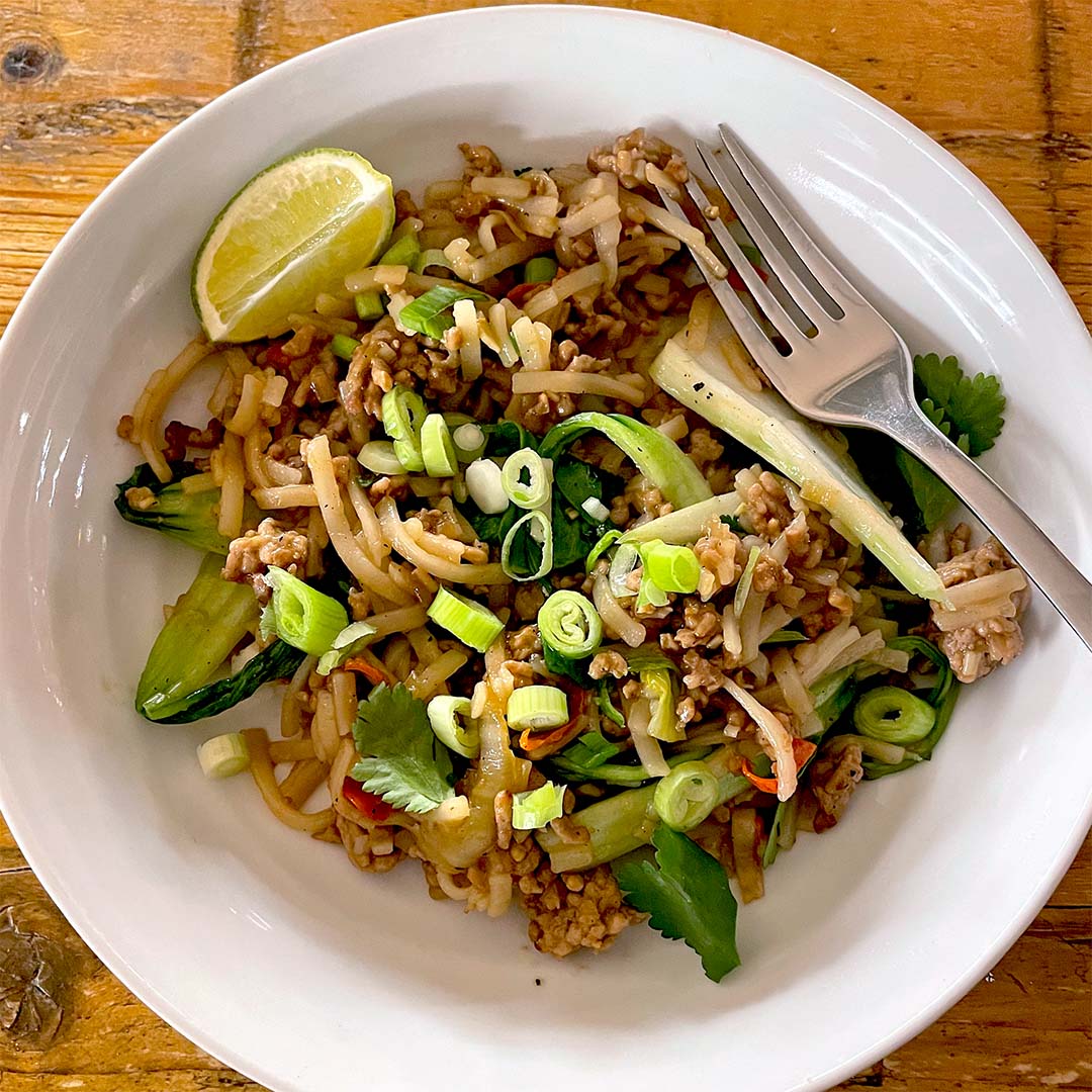 A white bowl filled with stir-fried noodles, minced meat, sliced spring onions, coriander, cucumber, and a lime wedge, with a fork resting on the edge of the bowl, on a wooden table.
