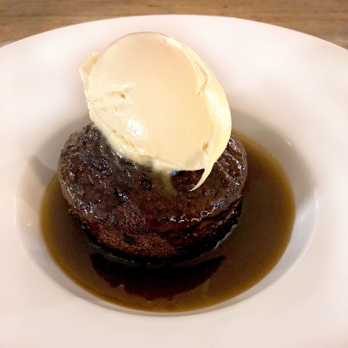 A dessert of sticky toffee pudding sits in a pool of toffee sauce on a white plate, topped with a scoop of vanilla ice cream.