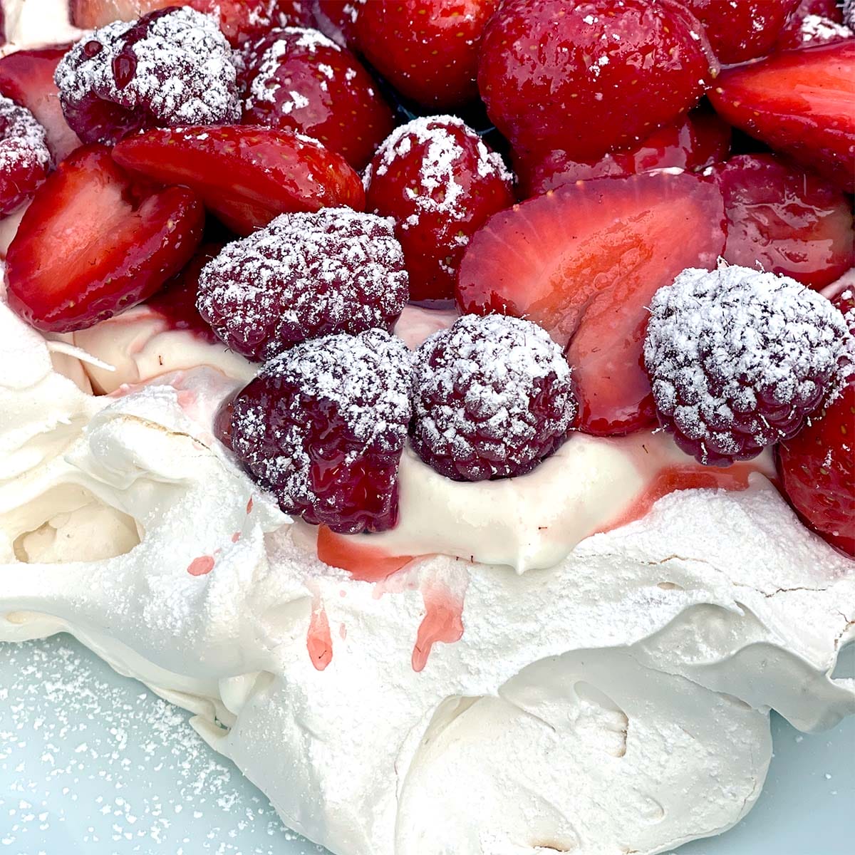 A close-up of a pavlova dessert topped with whipped cream, fresh strawberries, raspberries, and a dusting of icing sugar on a white plate.