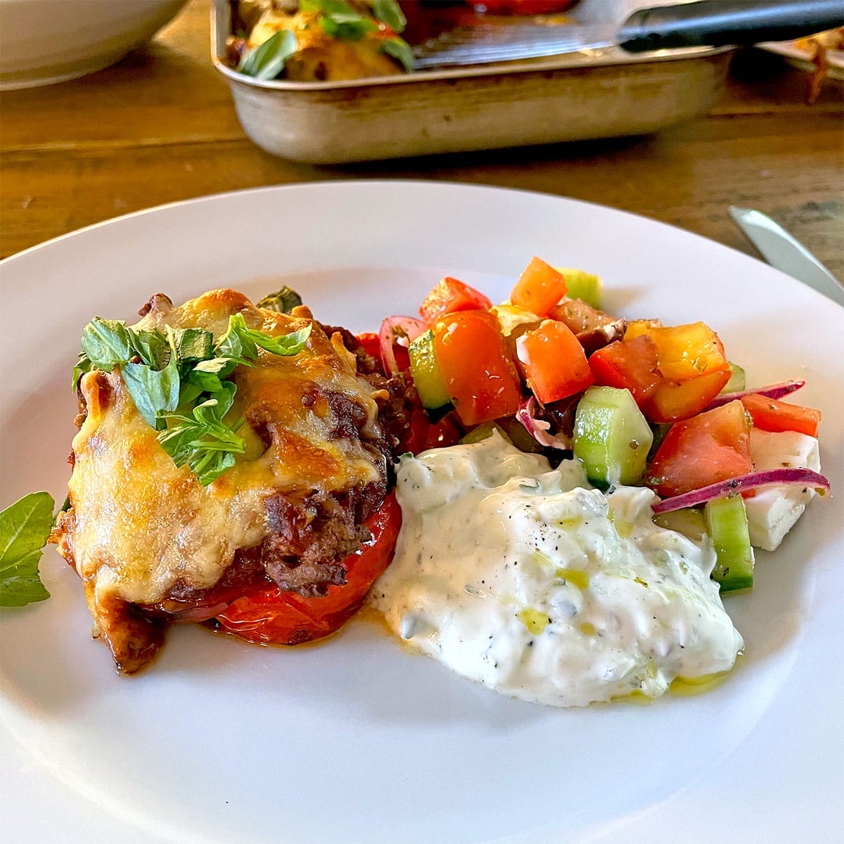 A white plate with a serving of baked dish topped with melted cheese and herbs, a scoop of creamy tzatziki, and a colourful Greek salad with tomatoes, cucumber, red onion, and feta cheese. A baking dish is visible in the background.