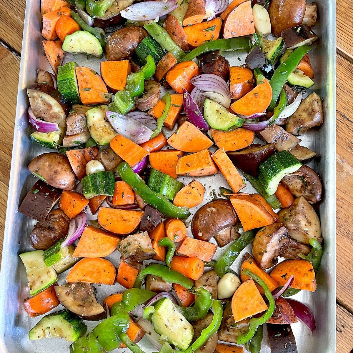 A baking tray filled with chopped mixed vegetables including sweet potatoes, courgette, aubergine, carrots, green pepper, red onion, mushrooms, and whole garlic cloves, ready for roasting.