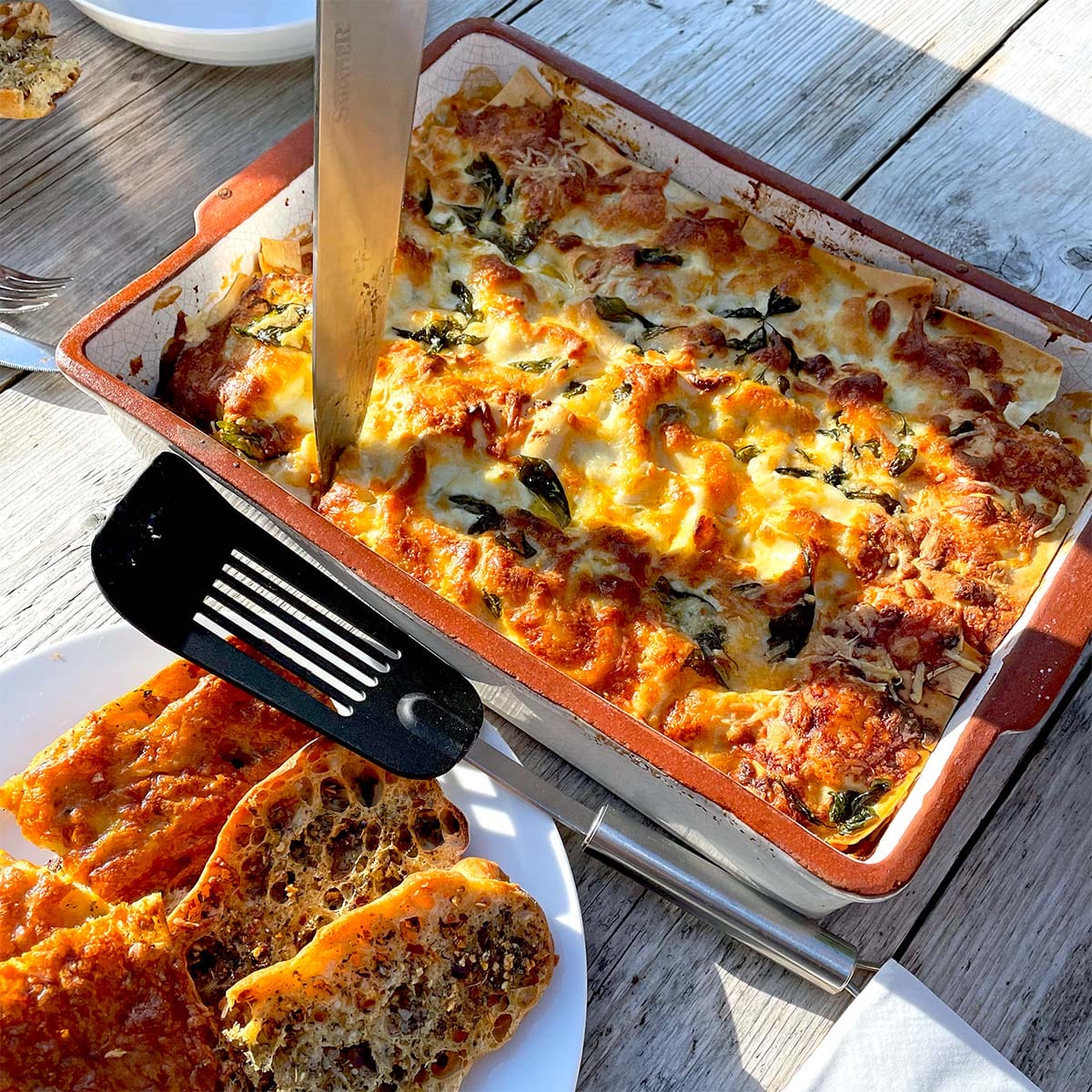 A baked lasagne in a rectangular dish with a knife inserted, surrounded by golden melted cheese and herbs. A plate with slices of crusty bread and a black spatula sits nearby on a sunlit wooden table.