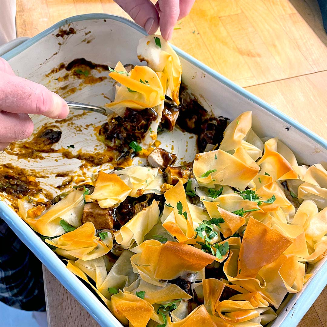 A person serves a portion of a baked dish with golden, crispy filo pastry on top, revealing a rich, dark filling underneath in a white rectangular baking dish. Parsley is sprinkled on the pastry.