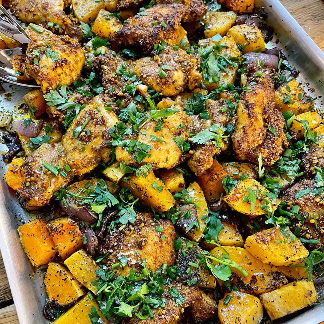 A tray of roasted chicken pieces and diced vegetables, including yellow courgette, onions, and herbs, garnished with chopped fresh parsley and spices. The dish appears golden-brown and flavourful.