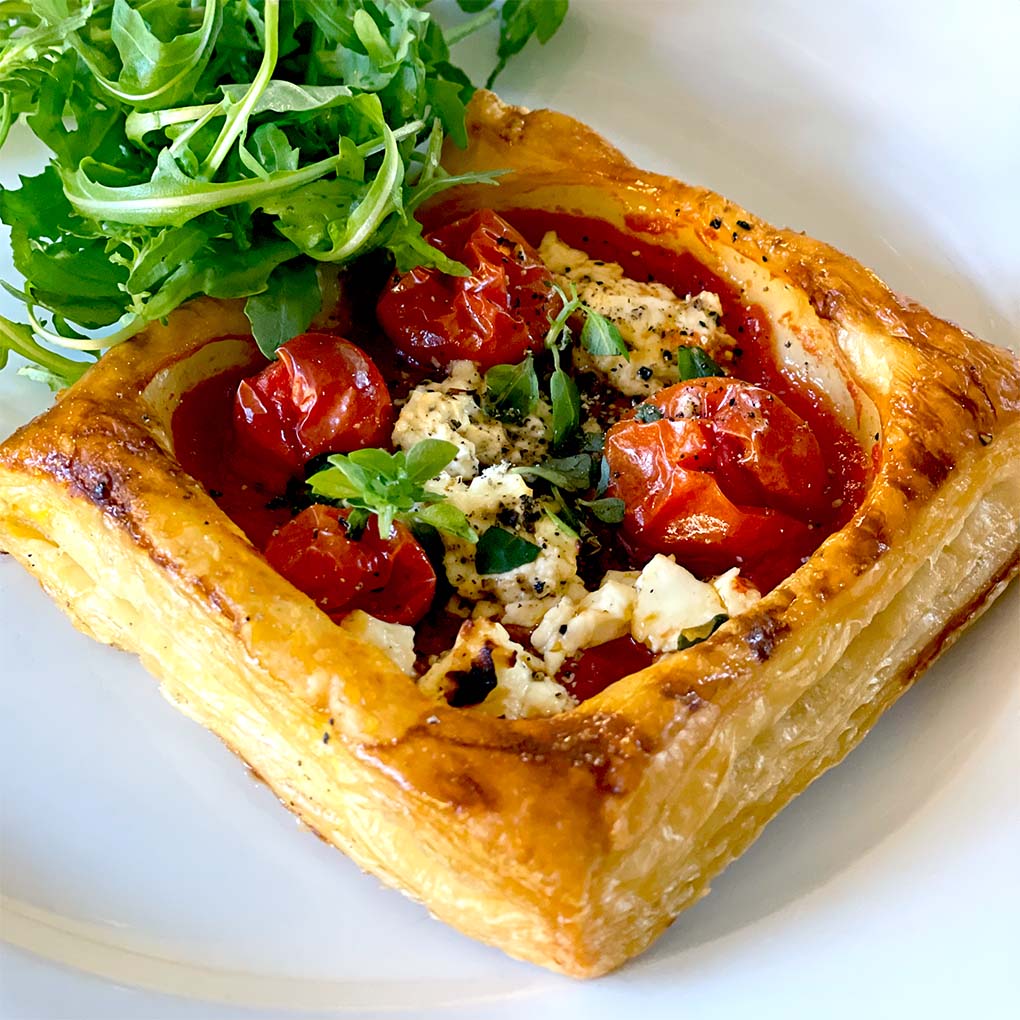 A golden puff pastry square filled with roasted cherry tomatoes, crumbled cheese, and herbs, served on a white plate with a side of fresh rocket.