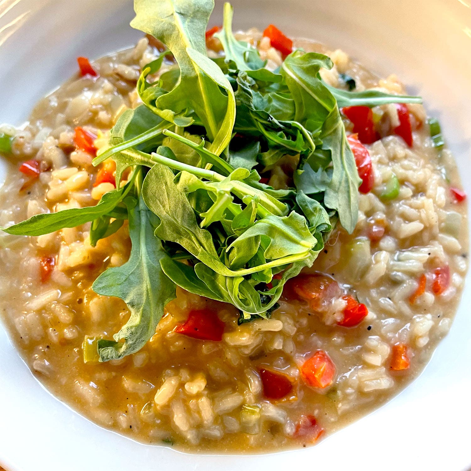 A close-up of creamy risotto with diced red peppers, garnished with a handful of fresh rocket, served in a white bowl.