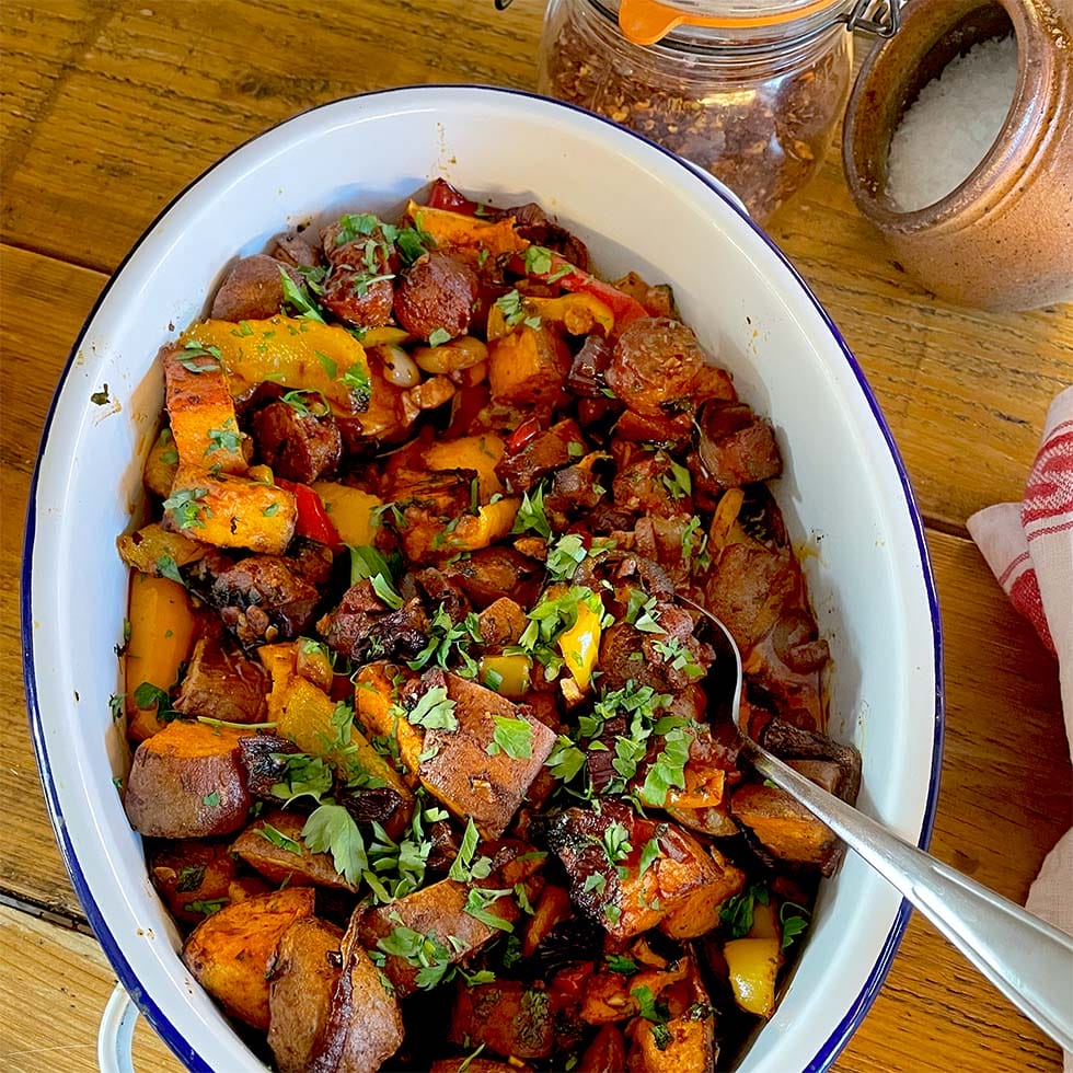 A white oval dish filled with a colourful roasted vegetable medley, including sweet potatoes, peppers, and mushrooms, garnished with chopped herbs. A spoon rests in the dish, and salt and chilli flakes are nearby.