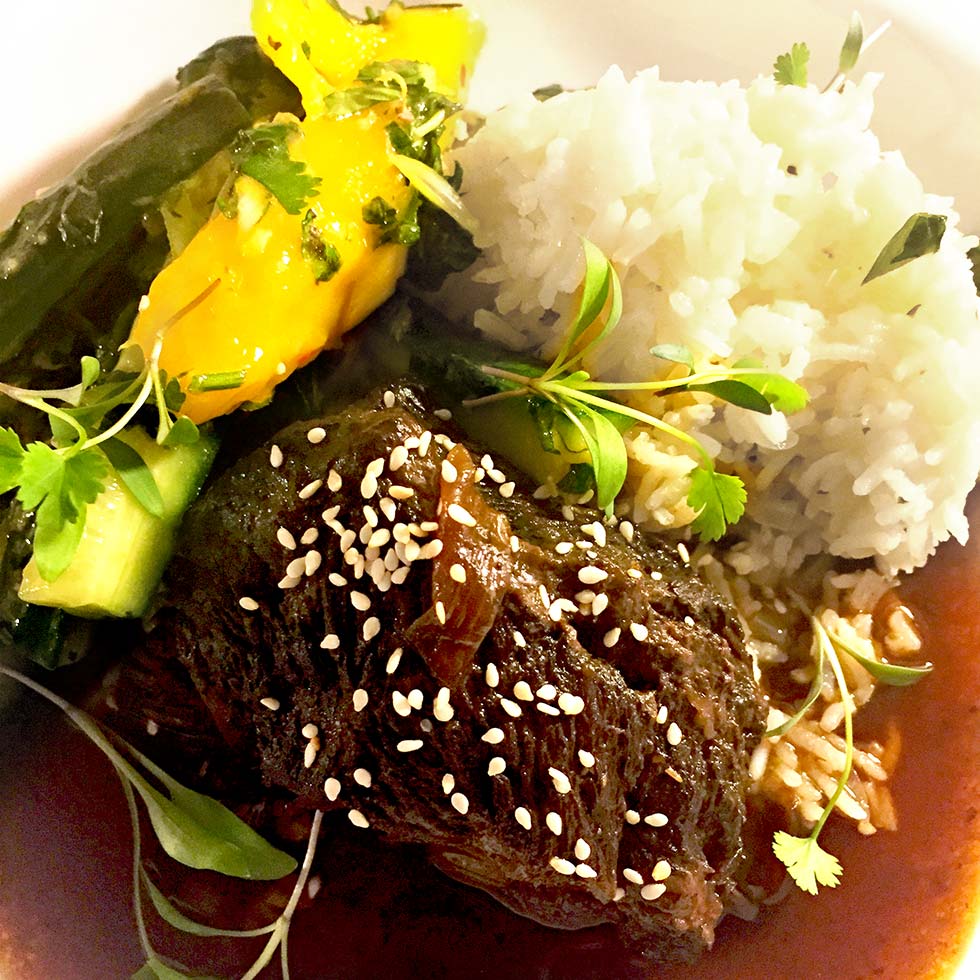 Braised beef topped with sesame seeds, served with white rice, sautéed green vegetables, and slices of yellow pepper, garnished with fresh herbs on a white plate.