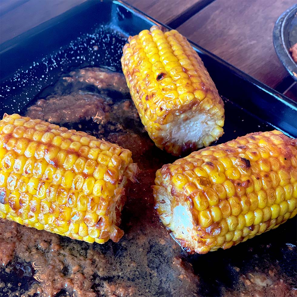 Three pieces of grilled corn on the cob, coated with seasoning and butter, sit on a black tray with melted butter and spices pooling around them.